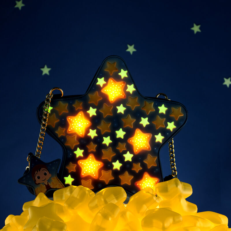 Star-shaped Loungefly La Luna Figural Crossbody sitting on a pile of gold stars against a nighttime sky background with the lights low to show how the stars on the bag light up and glow in the dark 
